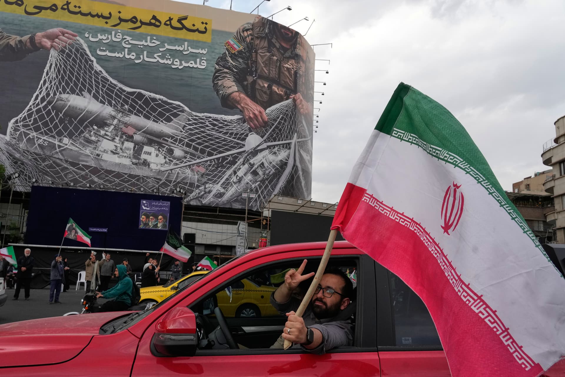 AP/Vahid Salemi A man flashes a victory sign as he carries an Iranian flag in front of an anti-U.S. billboard that reads in Farsi: 'The Strait of Hormuz will remain closed, The entire Persian Gulf is our hunting ground,' April 13, 2026.