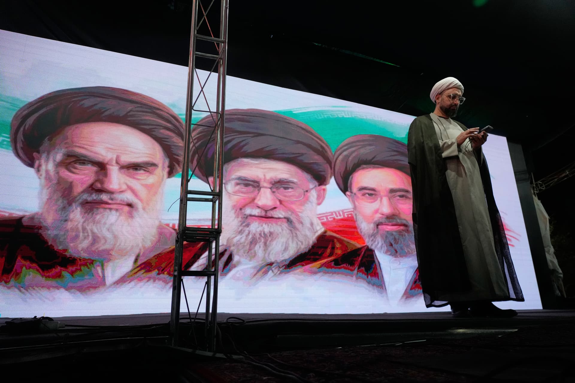 AP/Vahid Salemi A cleric checks his cell phone on stage in front of a screen displaying portraits of the late Iranian revolutionary founder Ayatollah Khomeini, left, late Supreme Leader Ayatollah Ali Khamenei, center, and Supreme Leader Ayatollah Mojtaba Khamenei, during a pro-government gathering in Tehran June 7, 2026.
