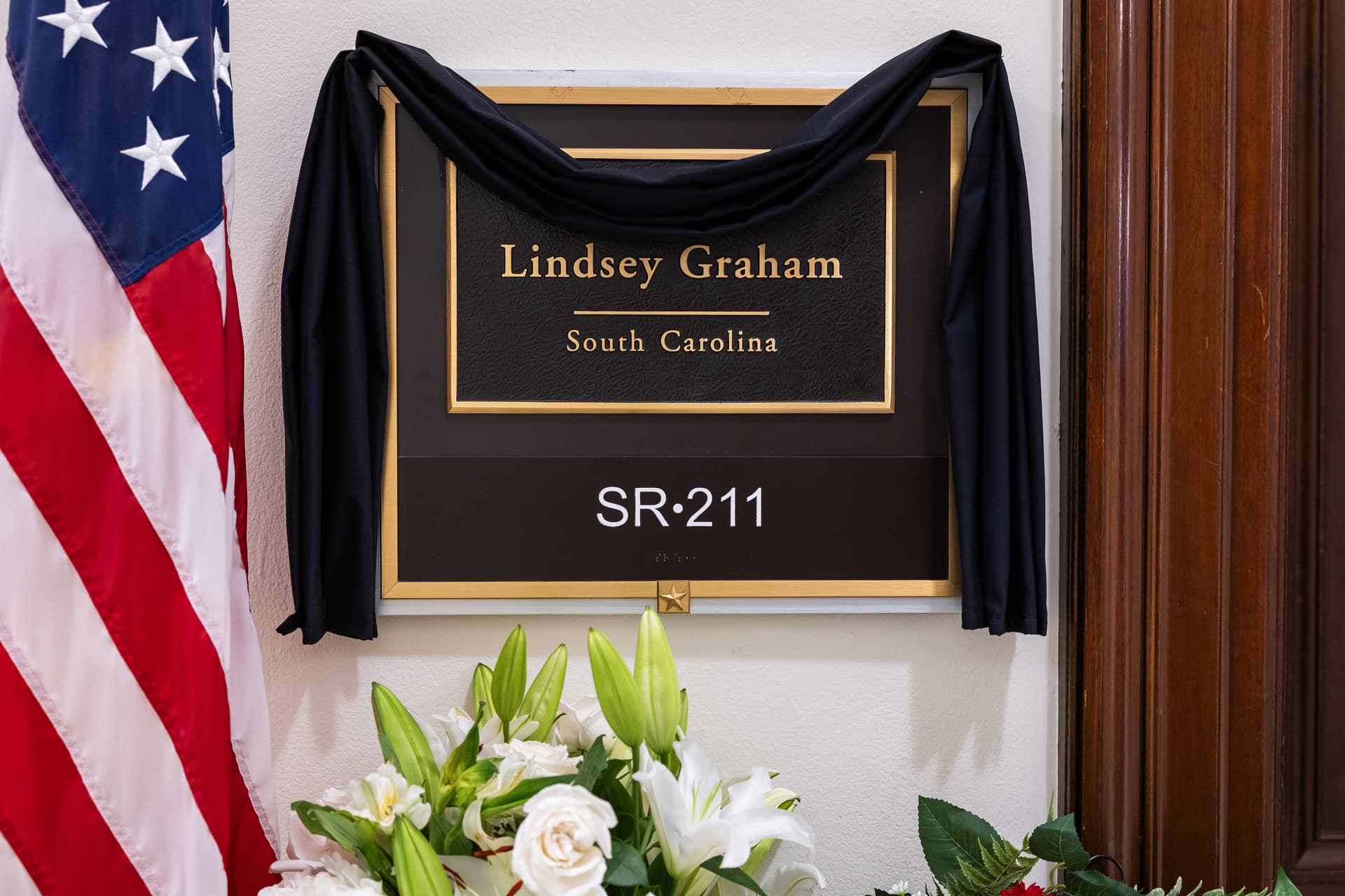 Finn Gomez/Getty Images Flowers surround the covered plaque outside the office of the late Senator Lindsey Graham at Russell Senate Office Building on July 14, 2026.